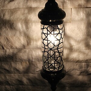 May include: A black metal and glass lantern with a geometric pattern. The lantern is lit and casting a warm glow.