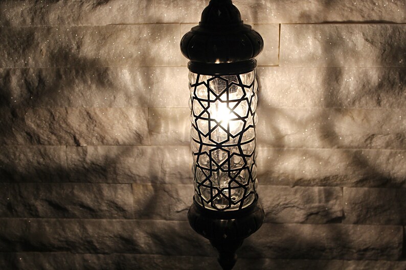 May include: A black metal and glass lantern with a geometric pattern. The lantern is lit and casting a warm glow.