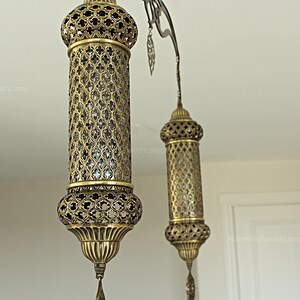 May include: Ornate brass pendant lights with intricate cut-out patterns. The lamps have a cylindrical shape with a decorative top and bottom, and a dangling tassel. The lamps are suspended from a curved metal fixture.