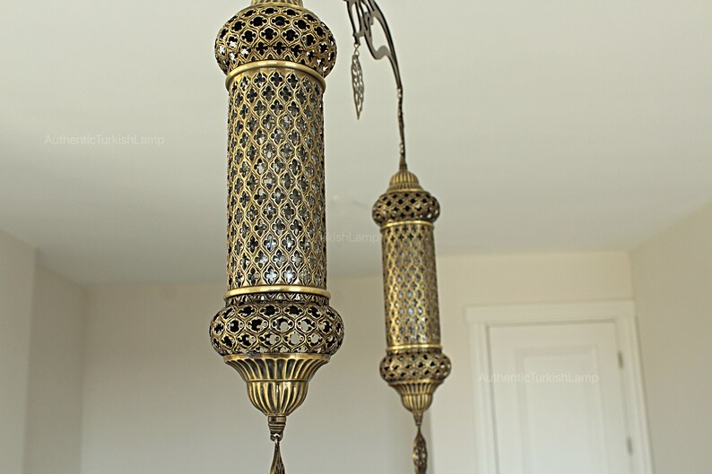 May include: Ornate brass pendant lights with intricate cut-out patterns. The lamps have a cylindrical shape with a decorative top and bottom, and a dangling tassel. The lamps are suspended from a curved metal fixture.