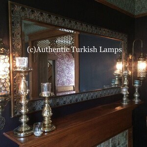 May include: A set of three ornate gold-toned metal candle holders with a decorative mirror in the background. The mirror has a carved wooden frame and a dark blue background. The text "(c) Authentic Turkish Lamps" is visible in the mirror reflection.