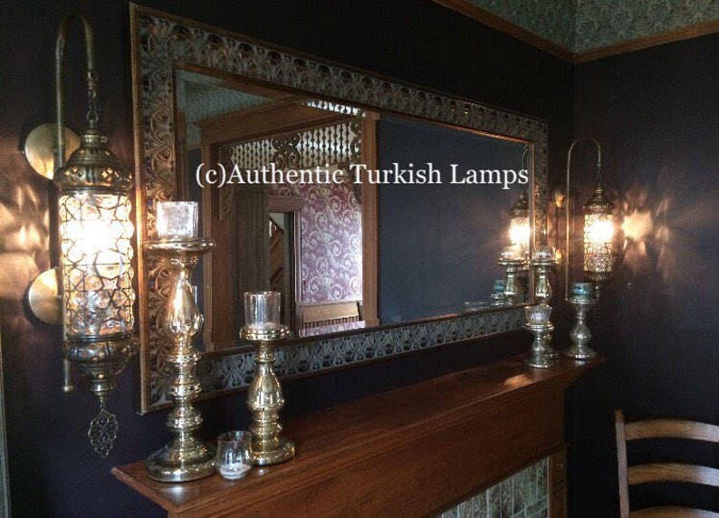May include: A set of three ornate gold-toned metal candle holders with a decorative mirror in the background. The mirror has a carved wooden frame and a dark blue background. The text "(c) Authentic Turkish Lamps" is visible in the mirror reflection.