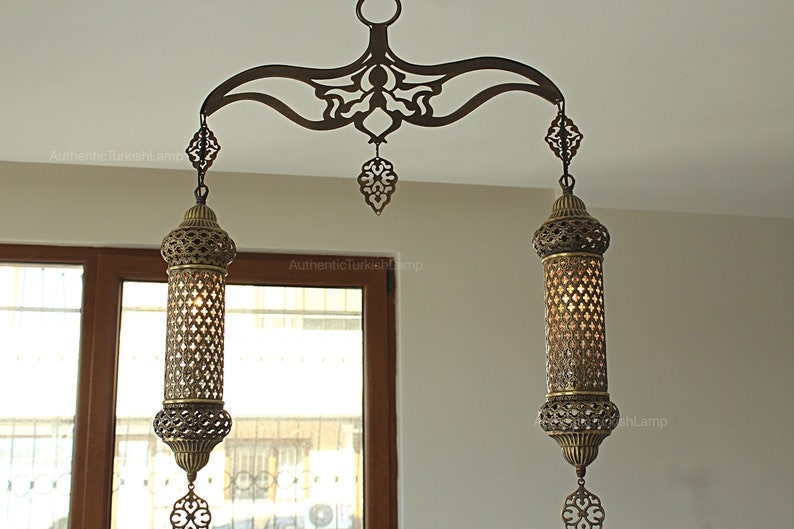 May include: An ornate bronze Turkish chandelier with two cylindrical lamps. The lamps have intricate cut-out patterns, suspended from a decorative arch. The design includes a central pendant and decorative elements.