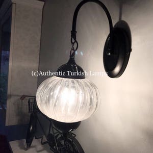 May include: A black wall sconce with a clear glass globe shade. The shade has a ribbed texture and is lit by a light bulb. The sconce is mounted on a wall with a white trim. (c) Authentic Turkish Lamps