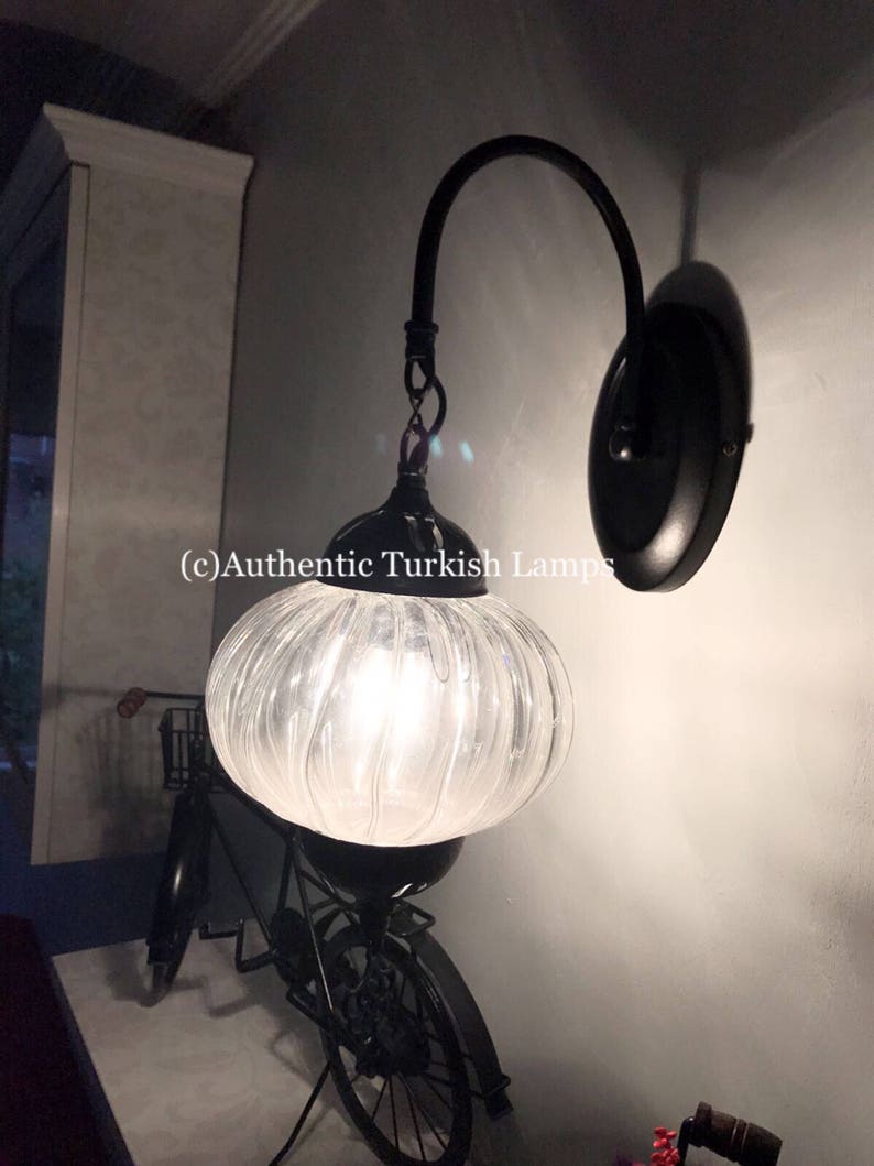 May include: A black wall sconce with a clear glass globe shade. The shade has a ribbed texture and is lit by a light bulb. The sconce is mounted on a wall with a white trim. (c) Authentic Turkish Lamps