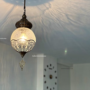 May include: A round, white glass pendant light with a decorative metal frame and a chain hanging from the ceiling. The light is on and casting a warm glow.
