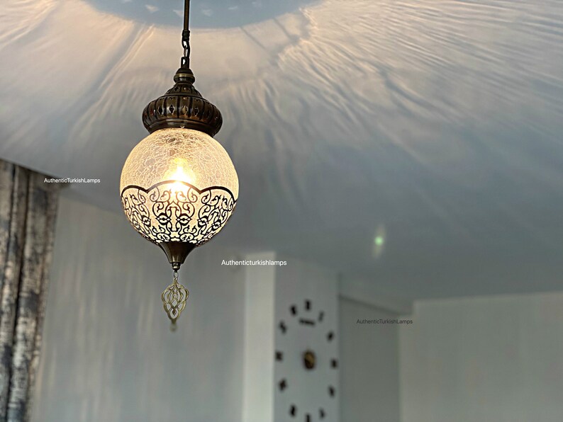May include: A round, white glass pendant light with a decorative metal frame and a chain hanging from the ceiling. The light is on and casting a warm glow.