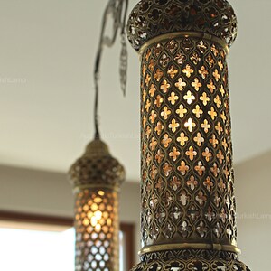 May include: Close-up of two ornate brass pendant lamps. The lamps have intricate cut-out patterns, casting a warm glow. The design is reminiscent of traditional Turkish lanterns, adding an exotic touch to any space.