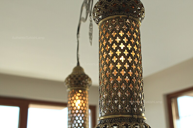 May include: Close-up of two ornate brass pendant lamps. The lamps have intricate cut-out patterns, casting a warm glow. The design is reminiscent of traditional Turkish lanterns, adding an exotic touch to any space.