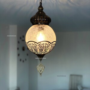 May include: A round, clear glass pendant light with a decorative metal frame and a chain hanging from the ceiling. The light is on and casting a warm glow.