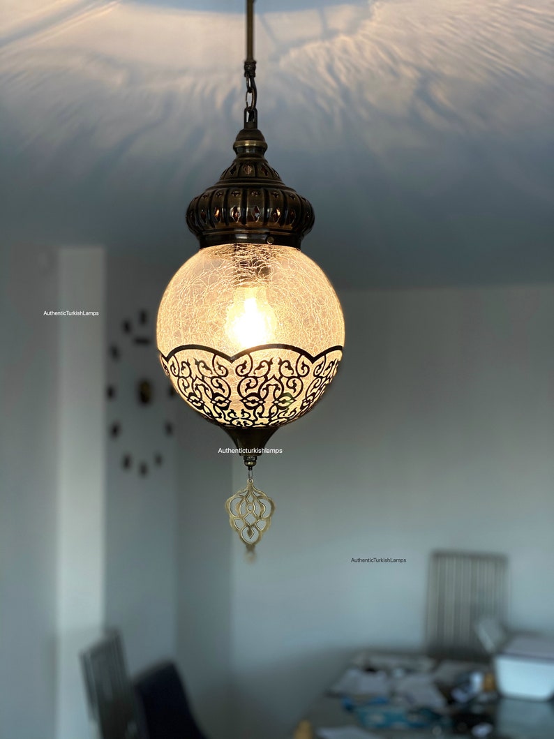 May include: A round, clear glass pendant light with a decorative metal frame and a chain hanging from the ceiling. The light is on and casting a warm glow.
