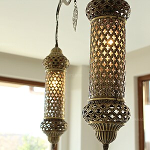 May include: Two ornate, antique-style pendant lights with a bronze finish hang from the ceiling. The cylindrical shades feature intricate cut-out patterns that allow light to filter through. The lights are suspended in front of a window.