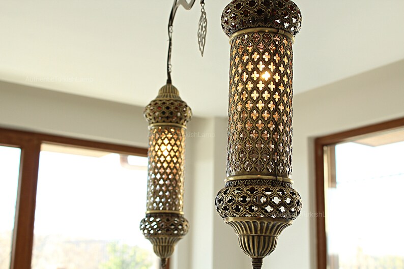May include: Two ornate, antique-style pendant lights with a bronze finish hang from the ceiling. The cylindrical shades feature intricate cut-out patterns that allow light to filter through. The lights are suspended in front of a window.