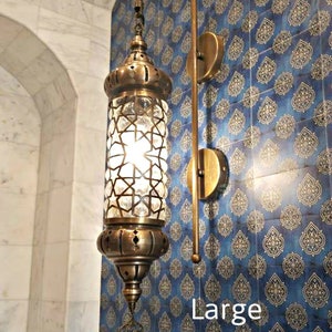 May include: A brass wall sconce with a clear glass shade featuring a geometric pattern. The sconce is mounted on a blue and gold patterned tile wall. The text "Large" is visible in the image.