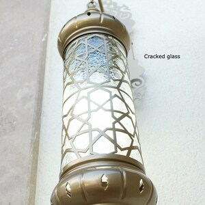May include: A gold-toned metal lantern with a clear glass panel featuring a geometric pattern. The glass panel has a cracked appearance.