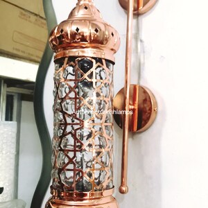 May include: A copper wall sconce with a clear glass shade featuring a geometric pattern. The sconce has a decorative finial and a chain hanging from the bottom. The sconce is mounted on a white wall. AuthenticTurkishLamps is printed on the wall.