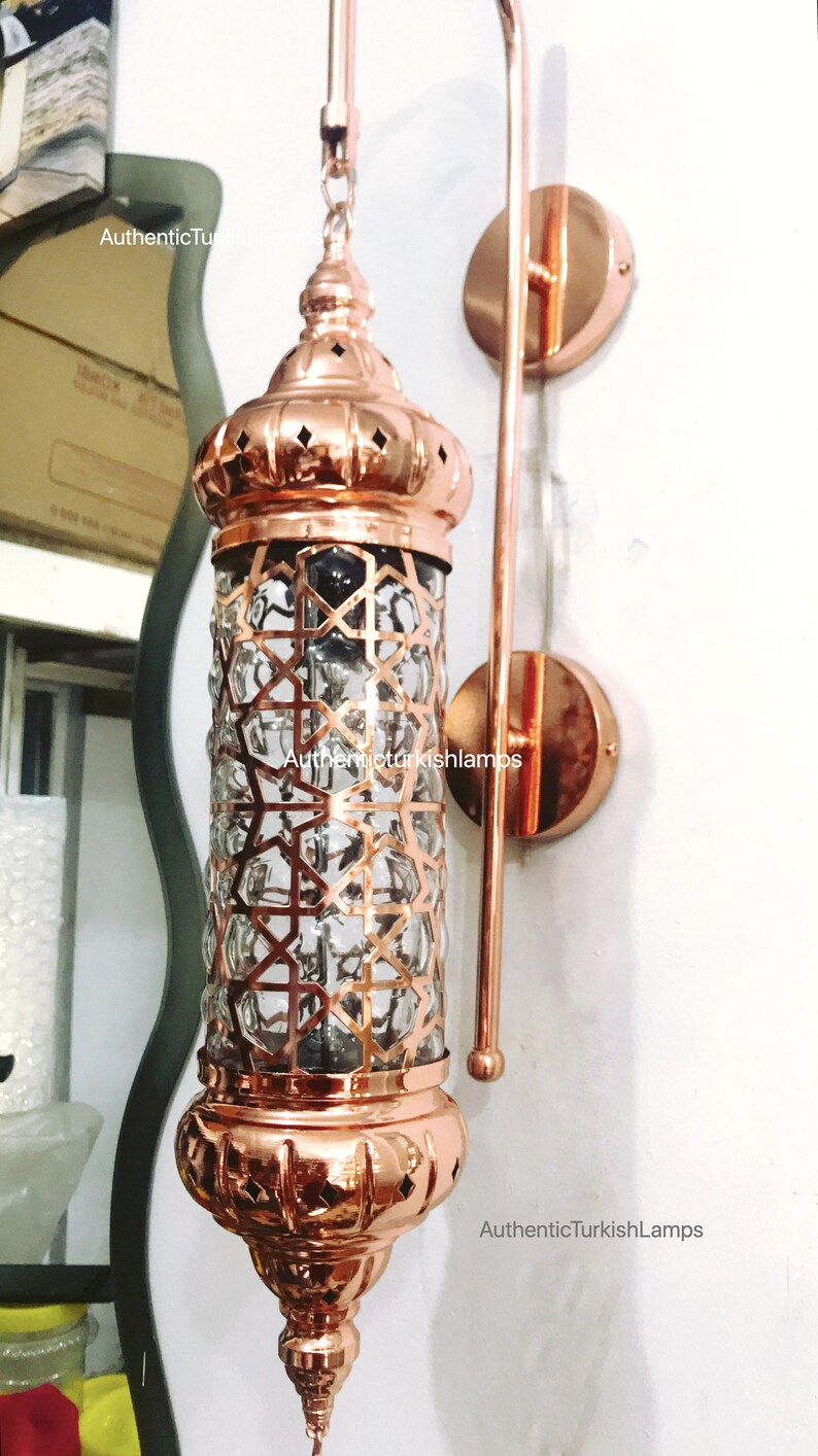 May include: A copper wall sconce with a clear glass shade featuring a geometric pattern. The sconce has a decorative finial and a chain hanging from the bottom. The sconce is mounted on a white wall. AuthenticTurkishLamps is printed on the wall.