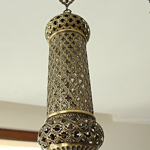 May include: An ornate hanging lamp with a cylindrical, openwork metal shade. The lamp has a detailed, geometric pattern and a warm, brassy finish. The design suggests a Middle Eastern or Turkish influence. The lamp is suspended from a chain.