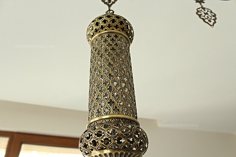 May include: An ornate hanging lamp with a cylindrical, openwork metal shade. The lamp has a detailed, geometric pattern and a warm, brassy finish. The design suggests a Middle Eastern or Turkish influence. The lamp is suspended from a chain.
