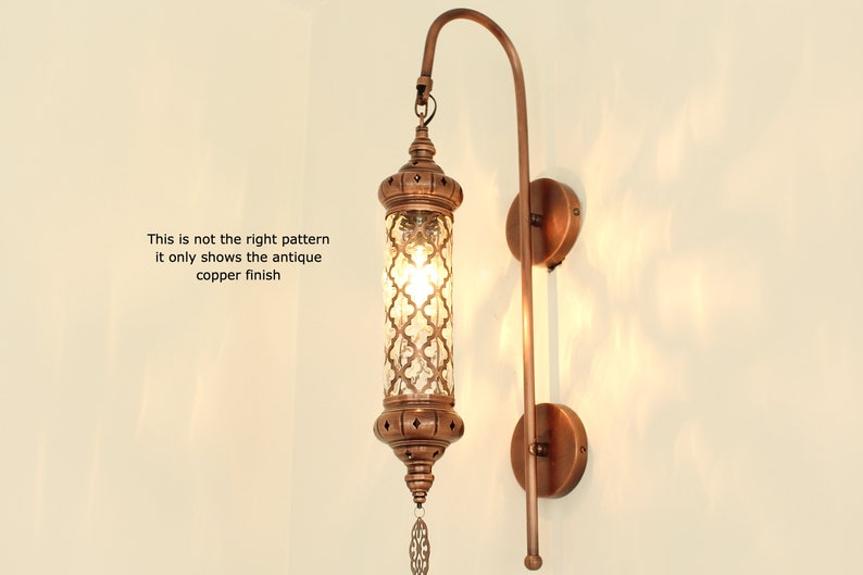 May include: An antique copper wall sconce with a cylindrical glass shade featuring a geometric pattern. The sconce has a curved arm and a round base.