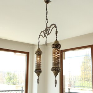 May include: An ornate, antique-style chandelier with two cylindrical lamps. The lamps are made of bronze-coloured metal with intricate cut-out patterns, hanging from a decorative fixture. The light fixtures are suspended from the ceiling.