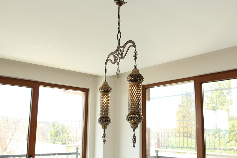 May include: An ornate, antique-style chandelier with two cylindrical lamps. The lamps are made of bronze-coloured metal with intricate cut-out patterns, hanging from a decorative fixture. The light fixtures are suspended from the ceiling.