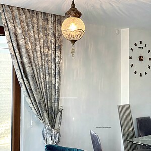 May include: A teal blue velvet sofa with a pink throw pillow and a white pillow with a blue butterfly design. A decorative hanging lamp with a clear glass globe and intricate metalwork hangs from the ceiling. The lamp is lit and casts a warm glow on the room. The room has a white wall with the text "AuthenticTurkishLamps" written on it.