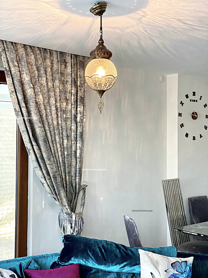 May include: A teal blue velvet sofa with a pink throw pillow and a white pillow with a blue butterfly design. A decorative hanging lamp with a clear glass globe and intricate metalwork hangs from the ceiling. The lamp is lit and casts a warm glow on the room. The room has a white wall with the text "AuthenticTurkishLamps" written on it.