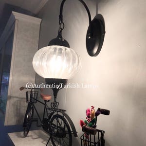 May include: A black wall sconce with a white glass shade. The sconce is mounted on a wall with a light blue background. The sconce is lit and casting a warm glow. The text (c) Authentic Turkish Lamps is visible in the image.