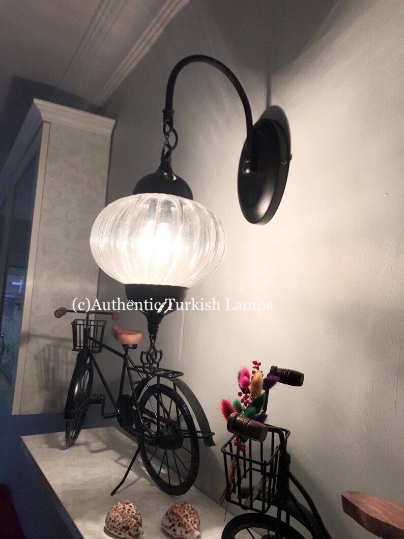 May include: A black wall sconce with a white glass shade. The sconce is mounted on a wall with a light blue background. The sconce is lit and casting a warm glow. The text (c) Authentic Turkish Lamps is visible in the image.