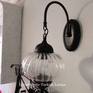 May include: A black metal wall sconce with a clear glass globe shade. The sconce has a curved arm and a round base. The glass shade has a ribbed texture. The sconce is mounted on a white wall. (c) Authentic Turkish Lamps