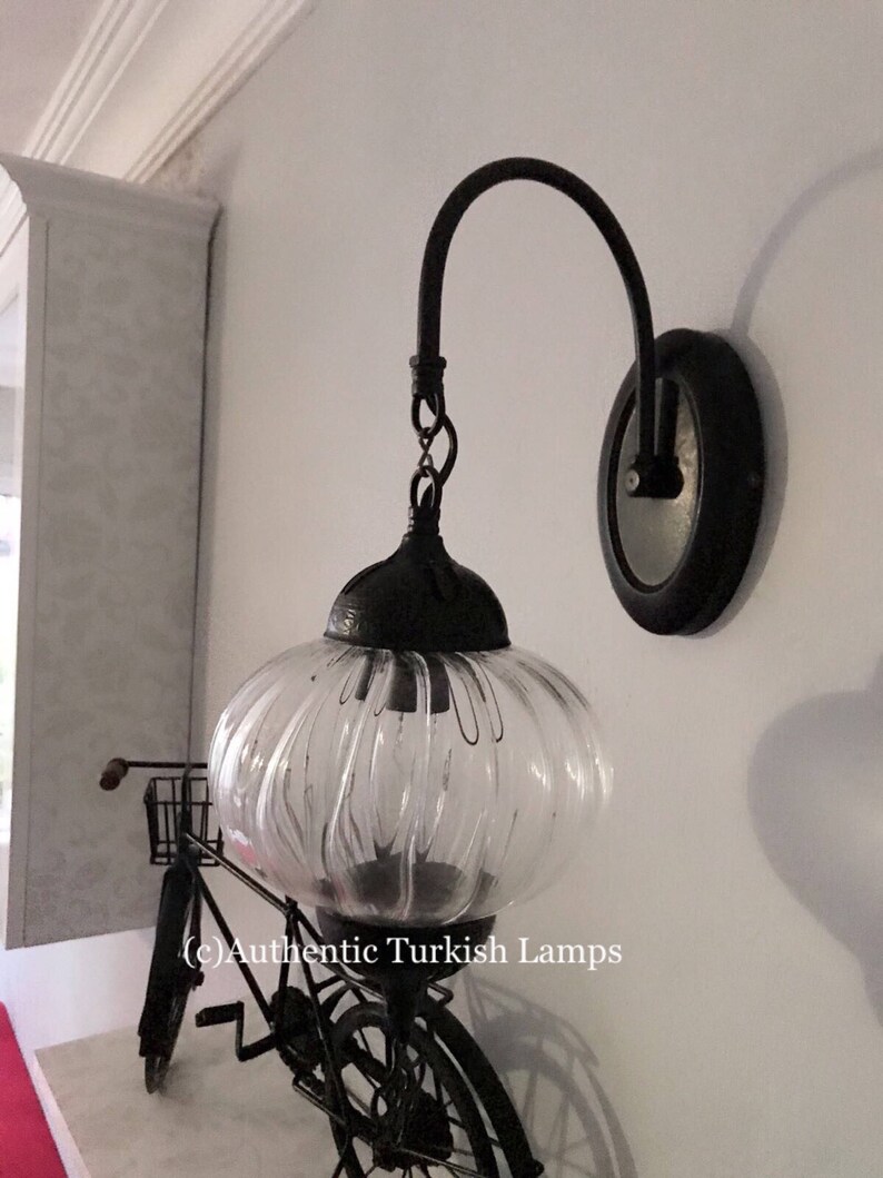 May include: A black metal wall sconce with a clear glass globe shade. The sconce has a curved arm and a round base. The glass shade has a ribbed texture. The sconce is mounted on a white wall. (c) Authentic Turkish Lamps