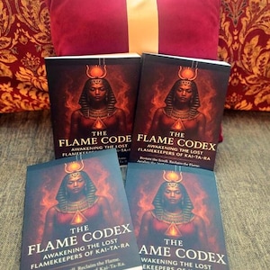 Puede incluir: Se muestran cuatro ejemplares del libro "The Flame Codex", con una ilustración de una figura egipcia en la portada. El título y subtítulo del libro, "Awakening the Lost Flamekeepers of Kai-Ta-Ra", se muestran de forma destacada. Un cojín burdeos con una raya dorada está en el fondo.