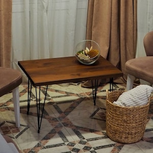 May include: A cozy living room scene featuring two brown armchairs, a dark wood coffee table with black hairpin legs, and a woven basket. The room is illuminated by pendant lights and framed by brown curtains and sheer white curtains.