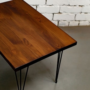 May include: A rectangular coffee table with a dark brown wooden top. The table has black hairpin legs. The wood grain is visible. The table is set against a white brick wall and a grey floor.