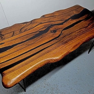 May include: A live edge coffee table with a natural wood top and black epoxy resin filling the cracks. The table has a unique, organic shape and is supported by thin black metal hairpin legs.
