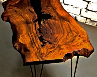 May include: Live edge coffee table with a unique, organic shape. The tabletop is crafted from a single piece of wood, showcasing rich brown and amber tones with a dark, black resin inlay. The table is supported by black metal hairpin legs.