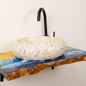 Può includere: Un lavabo in ceramica bianca con macchie nere si trova su un piano in legno con un inserto in resina epossidica blu e gialla. Un rubinetto nero è montato sul muro sopra il lavabo.