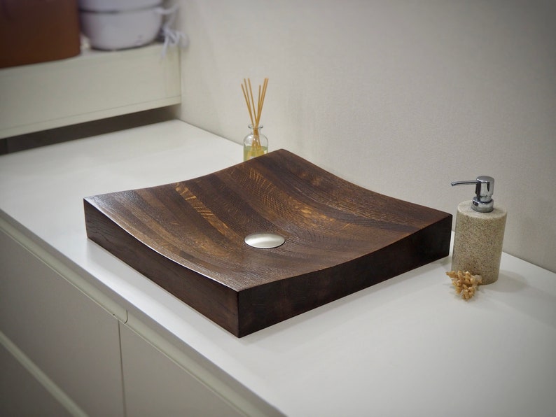 Peut inclure: Un lavabo de salle de bain en bois brun fonc&eacute; avec un design arrondi et incurv&eacute;. L'&eacute;vier a un bouchon de vidange argent&eacute; et est pos&eacute; sur un comptoir blanc.