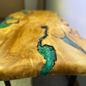 May include: A live edge wooden table with a natural, light brown finish. The table features a river of vibrant teal epoxy resin flowing through the center, creating a unique design. The table is supported by black metal legs.