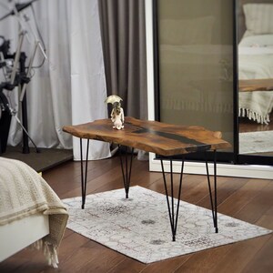 May include: A live edge wooden coffee table with a black epoxy resin inlay. The table has black hairpin legs and is sitting on a rug with a white background and brown accents.