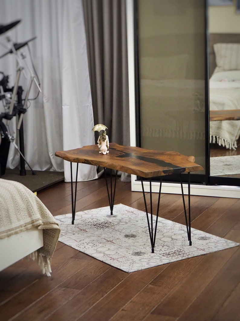 May include: A live edge wooden coffee table with a black epoxy resin inlay. The table has black hairpin legs and is sitting on a rug with a white background and brown accents.