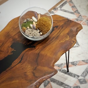 Puede incluir: Una mesa de café de madera con un diseño de borde vivo y una incrustación de resina epoxi negra. La mesa tiene un cuenco de vidrio en la parte superior lleno de musgo y flores secas.