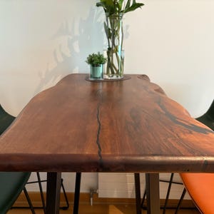 Peut inclure: Table en bois massif avec bords naturels et finition marron foncé. La table a une forme organique et repose sur des pieds en métal noir. Des chaises vertes et oranges entourent la table. Un vase de fleurs et une petite plante sont posés sur la table.
