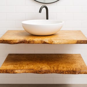 Peut inclure: Deux étagères flottantes en bois avec un bord naturel, d'une couleur brun doré chaleureux. Un lavabo rond blanc est posé sur l'étagère supérieure, avec un robinet noir. Les étagères sont fixées contre un mur carrelé blanc.