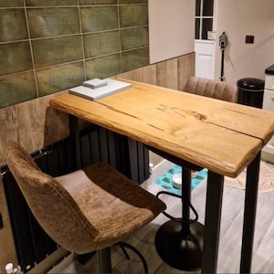 Puede incluir: Una mesa de bar de madera con un acabado de vetas naturales, sostenida por patas de metal negro. Una silla de bar giratoria de cuero marrón está colocada frente a la mesa. La mesa mide aproximadamente 122 cm de largo.