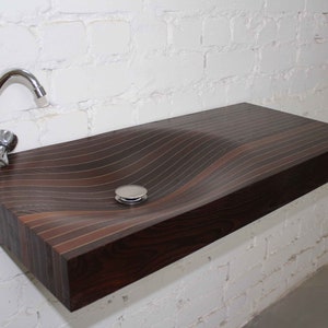 Könnte beinhalten: Ein dunkelbraunes Waschbecken aus Holz mit einem wellenförmigen Design und einem verchromten Wasserhahn. Das Waschbecken ist an einer weißen Backsteinwand montiert.