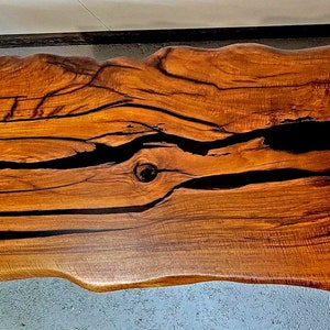 May include: A large, live edge wooden table top with a natural wood finish. The table top has a unique, irregular shape and features black epoxy resin filling in the cracks and knots of the wood.
