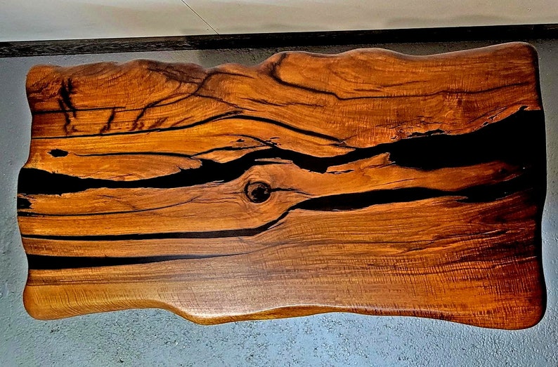 May include: A large, live edge wooden table top with a natural wood finish. The table top has a unique, irregular shape and features black epoxy resin filling in the cracks and knots of the wood.