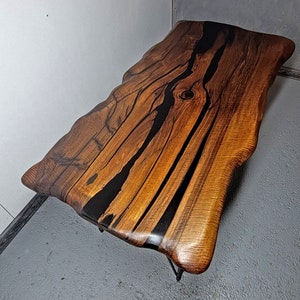 May include: A live edge coffee table with a natural wood top and black epoxy resin filling the cracks. The table has a unique, organic shape and is supported by a black metal frame.